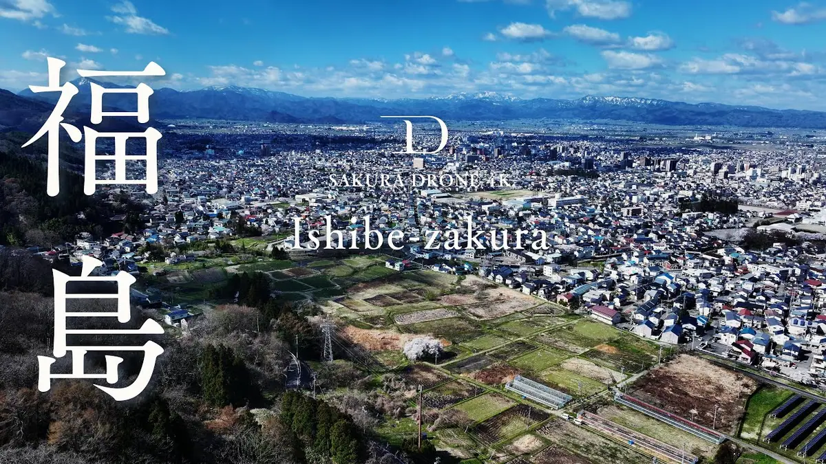 Aerial view of Ishibe-zakura town with fields and distant mountains; large white Japanese kanji Fukushima on the left and 'SAKURA DRONE AK'/'Ishibe-zakura' text overlayed in the center.